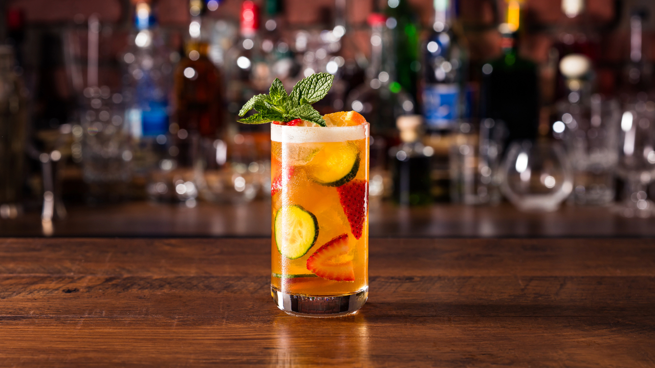 A Pimm's Cup cocktail on a bar in focus with various other bottles of liquor in the background