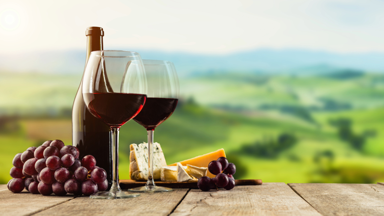 A bottle of wine, two glasses, a bundle of grapes, and blocks of cheese in front of a vineyard