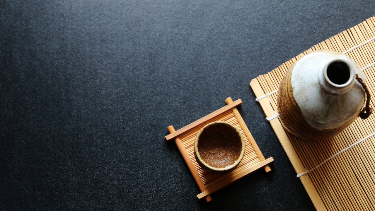 An overhead shot of a black table with a sake bottle, cup, and bamboo matt